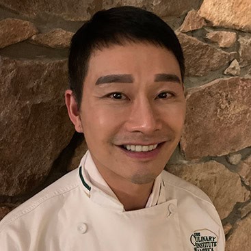 A man wearing a white chef's coat with "The Culinary Institute" logo stands against a stone wall. He is smiling at the camera.