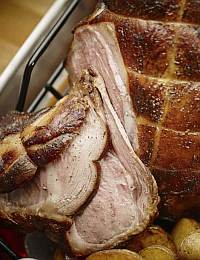 Close-up of a roasted ham partially sliced, revealing the juicy, cooked interior with a caramelized outer crust. The ham slices rest on whole roasted potatoes, and a meat rack is visible to the side.