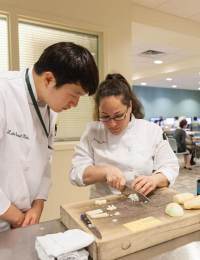 Two chefs in white uniforms are in a kitchen setting. One chef intently watches as the other chef chops vegetables on a cutting board. The background shows a busy kitchen with people working and various cooking equipment.