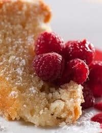 A slice of crumbly cake dusted with powdered sugar is served with a handful of vibrant red raspberries on a white plate. The cake appears moist and has a golden-brown crust, creating an inviting and appetizing dessert presentation.