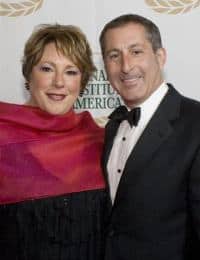 A woman in a red shawl and a man in formal attire, including a black tuxedo, stand close together and smile at the camera. The background features a patterned banner with laurels near the top.