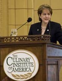 A person stands behind a podium with "The Culinary Institute of America" displayed on the front. They are speaking into a microphone inside a room with yellow walls. The person is wearing a dark blazer and gesturing with their hand.