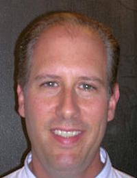 A man with short, light brown hair and a fair complexion smiles while facing the camera. He is wearing a light-colored shirt and is photographed against a dark background.