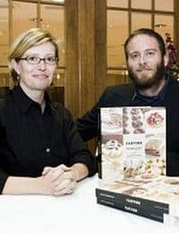 Two individuals, a woman with short blond hair and glasses, and a bearded man, are sitting at a table with stacked cookbooks titled "Tartine" in front of them. They appear to be at a book signing or promotional event in a warmly lit indoor setting.