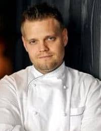 A man with short, light hair and a trimmed beard is wearing a white chef's jacket. He is standing against a dark background with a slight smile on his face. He has his arms crossed casually in front of him.