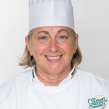 A person wearing a white chef's uniform and tall chef hat, smiling at the camera. The uniform has the logo of The Culinary Institute of America on it. The individual has short hair and appears to be standing against a plain white background.