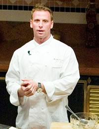 A chef wearing a white uniform stands in a kitchen. He is facing forward and appears to be speaking. There is a bowl with some ingredients on the counter in front of him. The background shows kitchen cabinets and a decorative tiled wall.