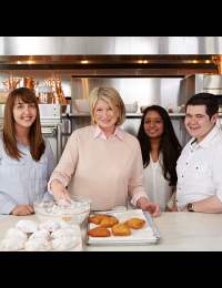 Four individuals stand in a kitchen setting. The person in the center, smiling, is preparing food with a bowl and a tray of pastries. The other three people, two women and a man, are also smiling and standing beside the central figure.