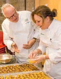 Two chefs in white uniforms are preparing food together in a kitchen. One chef, an older man with white hair and glasses, looks on and talks while the other, a woman, sprinkles seasoning onto a tray of food items. They appear to be enjoying the activity.