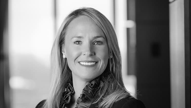 A black-and-white photograph shows a smiling woman with long, straight hair. She is wearing a dark blazer over a ruffled top. The background is blurred, featuring large windows that allow natural light to enter.