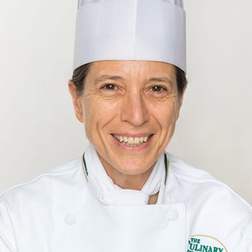 A chef smiling at the camera, wearing a white chef's uniform and tall white chef's hat. The uniform has a logo with the text, "The Culinary." The background is plain and light-colored.