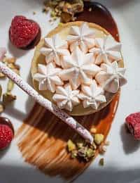 A small dessert features cream-colored meringue stars on top, set on a plate with a smear of chocolate sauce. Fresh raspberries and chopped nuts surround the dessert, along with a decorative swirled stick.