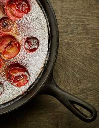 A cast iron skillet containing a baked dish with halved plums dusted with powdered sugar, placed on a rustic wooden surface. The plums are baked into a golden-brown batter, creating a visually appealing dessert.