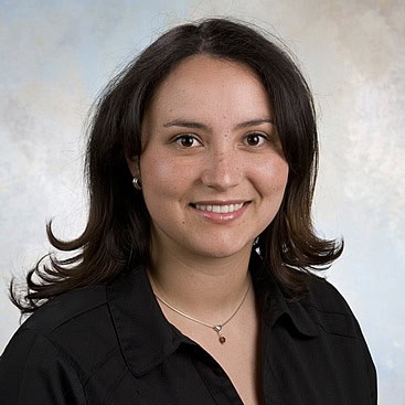 A woman with shoulder-length brown hair, wearing a black blouse and a necklace, is smiling at the camera against a light-colored background.