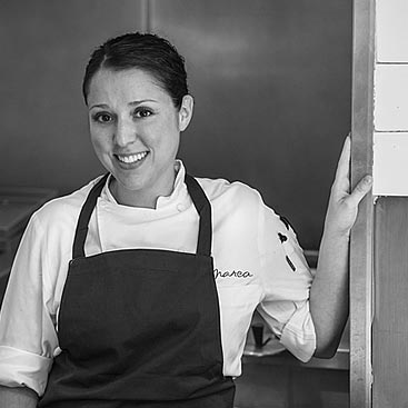 A smiling person in a white chef's coat and dark apron stands in a kitchen, leaning against a wall. The name "Marea" is embroidered on the chef's coat. The person has short dark hair and is positioned slightly to the left in the grayscale image.