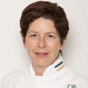 A person with short brown hair and a light complexion is wearing a white chef's coat with green and tan stripes on the collar. They are smiling slightly and looking directly at the camera against a plain white background.