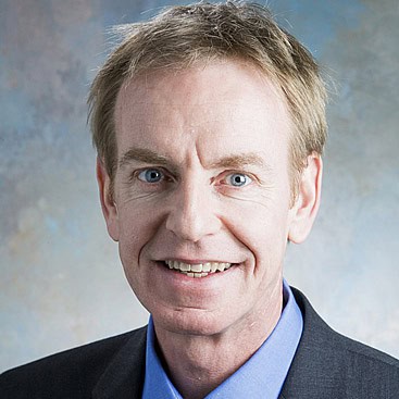 A man with short, light brown hair, blue eyes, and fair skin is smiling. He is wearing a blue dress shirt and a dark suit jacket. The background is softly blurred with shades of blue and gray.