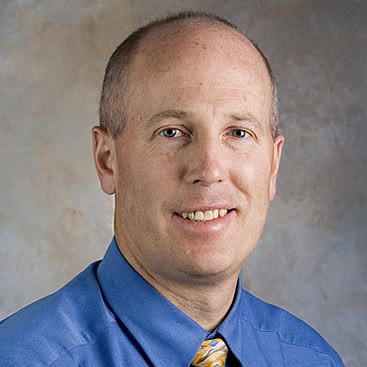 A middle-aged man with a receding hairline smiles at the camera. He is wearing a blue shirt and a yellow patterned tie. The background is neutral with gray and beige tones.