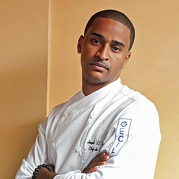 A man wearing a white chef's coat with the name "The Cecil" on the sleeve stands against a beige background, his arms crossed. He has short hair and a serious expression, with a black beaded bracelet on his left wrist.