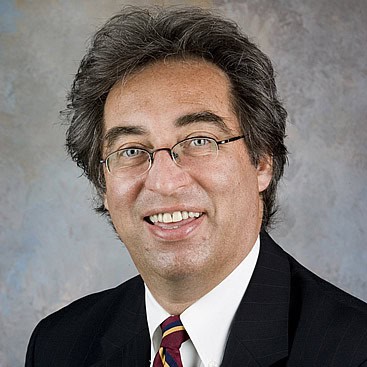 A man with gray, slightly wavy hair, wearing glasses, a striped tie in shades of red and yellow, and a dark suit with a white shirt, is smiling. The background is a neutral, textured backdrop.