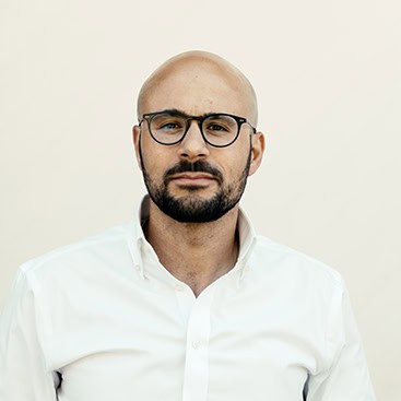 A bald man with a beard and mustache is wearing black-rimmed glasses and a white button-up shirt. He is looking directly at the camera against a plain off-white background.