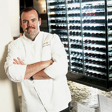 A chef with a mustache stands confidently with arms crossed in front of a wine rack filled with bottles. He is wearing a white chef's coat and smiling slightly. The setting appears to be a modern kitchen or restaurant with well-lit shelves displaying wine bottles.