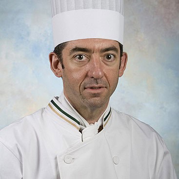 A man wearing a white chef's hat and a white chef's coat is facing the camera. The background is a soft blend of blue and beige hues. The chef's coat has a double-breasted design, and the collar has a subtle green and beige trim.