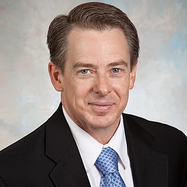 A man with short, neatly combed brown hair wearing a black suit jacket, white dress shirt, and blue patterned tie. He is smiling slightly against a neutral, cloud-like background.