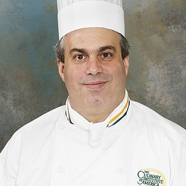 A professional chef wearing a white chef's coat and tall white chef hat. The coat has a logo that reads "The Culinary Institute of America." The chef has a neutral expression and is posing against a mottled blue and gray background.