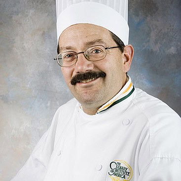 A middle-aged man with a mustache and glasses is wearing a white chef's hat and chef's coat. The coat features an embroidered logo with the text "The Culinary Institute of America." The background is a textured mix of blue and gray tones.