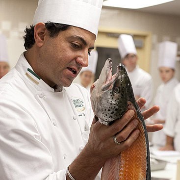 A chef in a white uniform and hat is holding and examining a large fish, likely a salmon, in a professional kitchen. Several other chefs in similar white uniforms and hats are visible in the background.