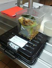 A clear plastic container filled with food waste sits on a scale in a commercial kitchen. The scale has a white label with writing. A red notepad is visible in the background on a stainless steel countertop.