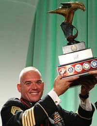 A man in a formal military uniform with several medals smiles as he proudly lifts a large trophy adorned with a golden eagle. The background features a green and white gradient.