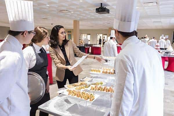 A woman in a beige blazer is talking to three chefs in white uniforms and tall chef hats, pointing at a table with various plated dishes. The setting appears to be a culinary classroom where chefs earn and learn, with other chefs working diligently in the background.