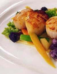 Seared scallops served on a plate with vibrant vegetables including yellow carrots, broccoli, and purple cauliflower. The dish is artistically plated to highlight the colors and textures of the ingredients.