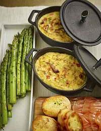 A wooden surface displays two cast-iron pots filled with a baked casserole, garnished with green herbs. Beside the pots are neatly arranged asparagus spears, slices of toasted bread, and smoked salmon, offering a variety of textures and colors.