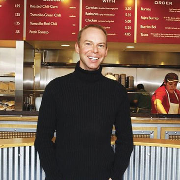 A person in a black turtleneck sweater is standing and smiling in front of a restaurant counter. The menu board in the background lists various food items and prices. A worker, in a red shirt and apron, is visible behind the counter.