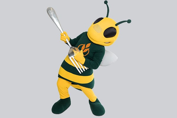 A bee mascot with black and yellow stripes, large black eyes, and a pair of green antennae, wearing a green shirt with a wheat logo. The mascot holds an oversized silver fork, standing on a light grey background.