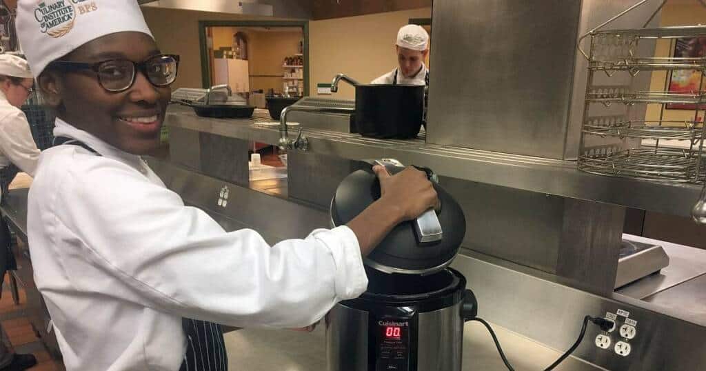 A person wearing a white chef's hat and coat is smiling while using a pressure cooker in a professional kitchen. Another person in a similar uniform is in the background, focusing on a pot on the stove. Stainless steel kitchen equipment surrounds them.