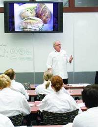 A chef in a white uniform is giving a presentation in a classroom. He is standing in front of a whiteboard and television screen displaying images of prepared dishes. Students, also in white uniforms, are seated and facing him, attentively listening.