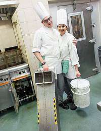 Two chefs in white uniforms and tall hats stand together in a commercial kitchen, smiling. The male chef on the left holds a large metal kitchen tool, and the female chef on the right holds a white bucket. They are standing in front of a stainless steel range and oven.