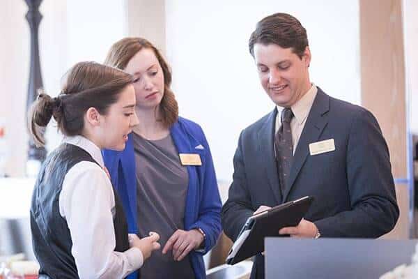 Three people in a professional setting are gathered and having a discussion. One person, wearing a vest, appears to be listening attentively. Another, in a blue blazer, looks on with interest. The third, a man in a suit, is showing something on a tablet—a clear opportunity to earn and learn together.