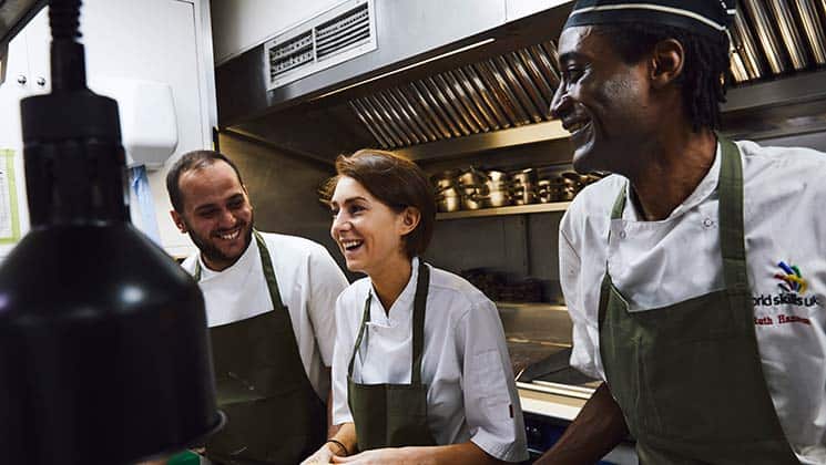 A group of three chefs in white uniforms and green aprons stand in a professional kitchen, smiling and engaging in conversation. The kitchen is equipped with stainless steel appliances and cookware.