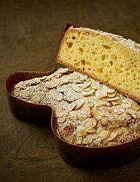 A sliced Italian Easter cake, known as Colomba, is shown on a wooden surface. The cake is light and yellow inside with a fluffy texture, while the top is adorned with almond slices and dusted with powdered sugar. It is shaped like a dove.