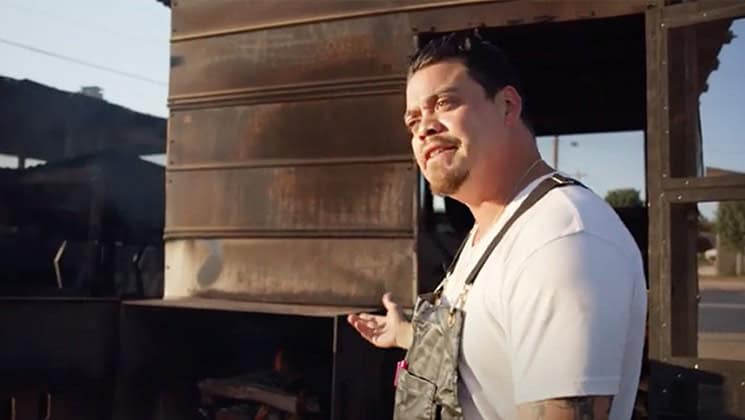 A man wearing a white shirt and denim overalls stands outside next to a large, rusty metal structure. He gestures with his right hand. The background shows an outdoor setting with trees and a clear sky.