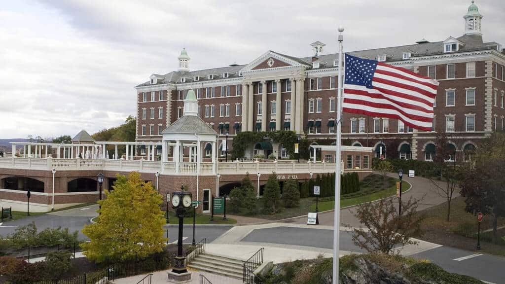 A large brick building with white columns and multiple windows stands on a landscaped campus. An American flag flies prominently in the foreground. Trees, pathways, a clock, and well-maintained gardens surround the building. Overcast sky above.