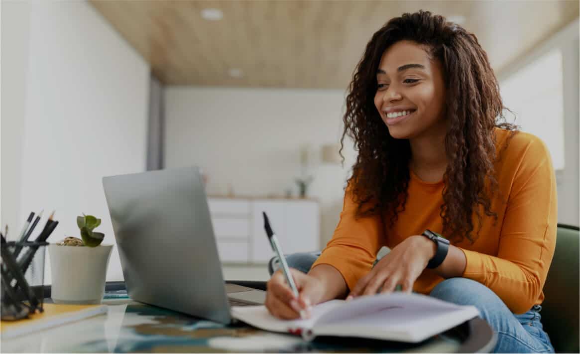 A woman with long curly hair is sitting at a desk and smiling while looking at a laptop. She is wearing an orange shirt and a smartwatch, and she is writing in a notebook with a black pen. A pencil holder and a small potted plant are on the desk beside her.