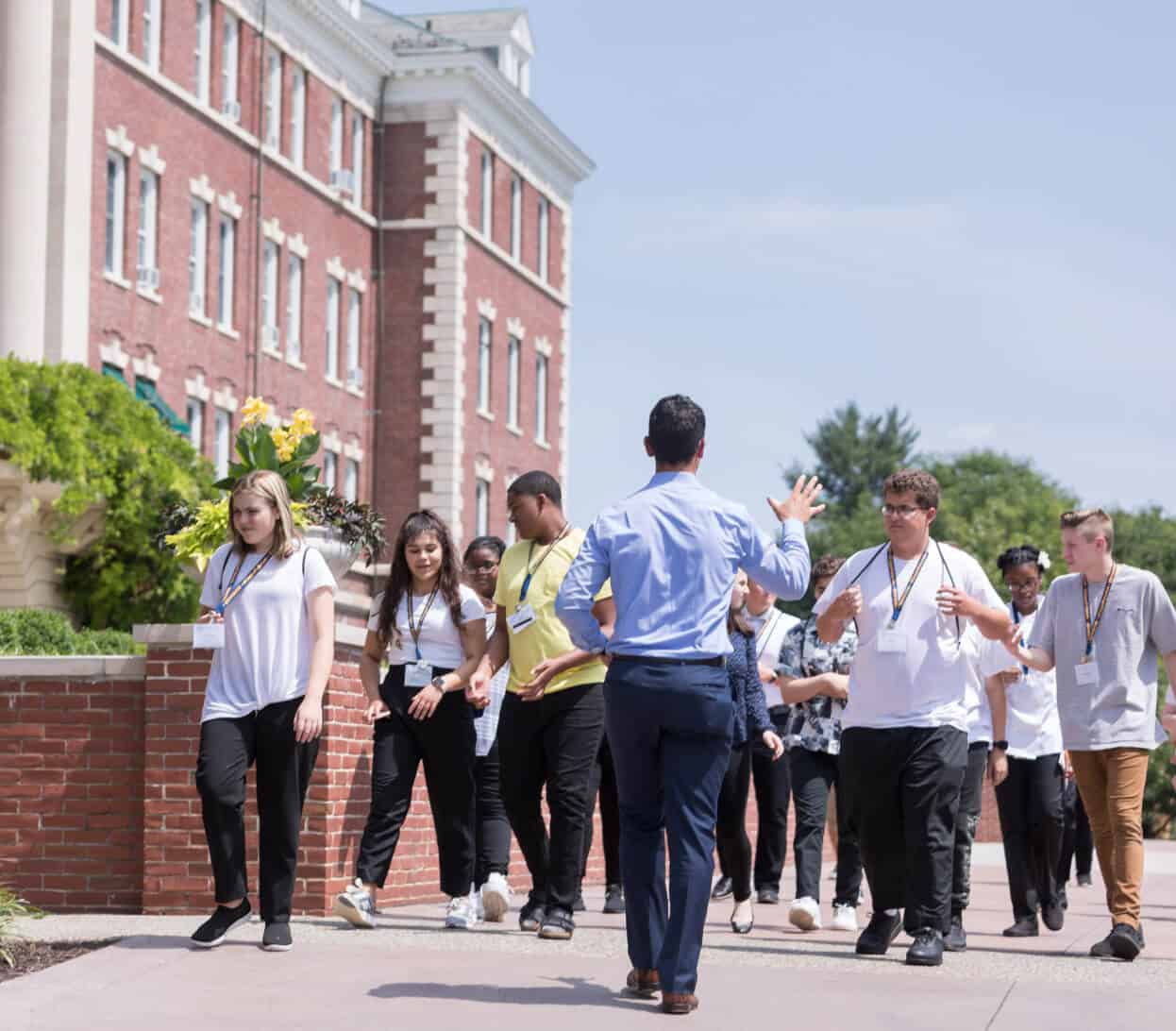 A group of students follow an instructor during a campus tour. They are walking on a paved pathway beside a large red-brick building with white-trimmed windows and green foliage. The instructor is gesturing with his right hand while talking to the students.