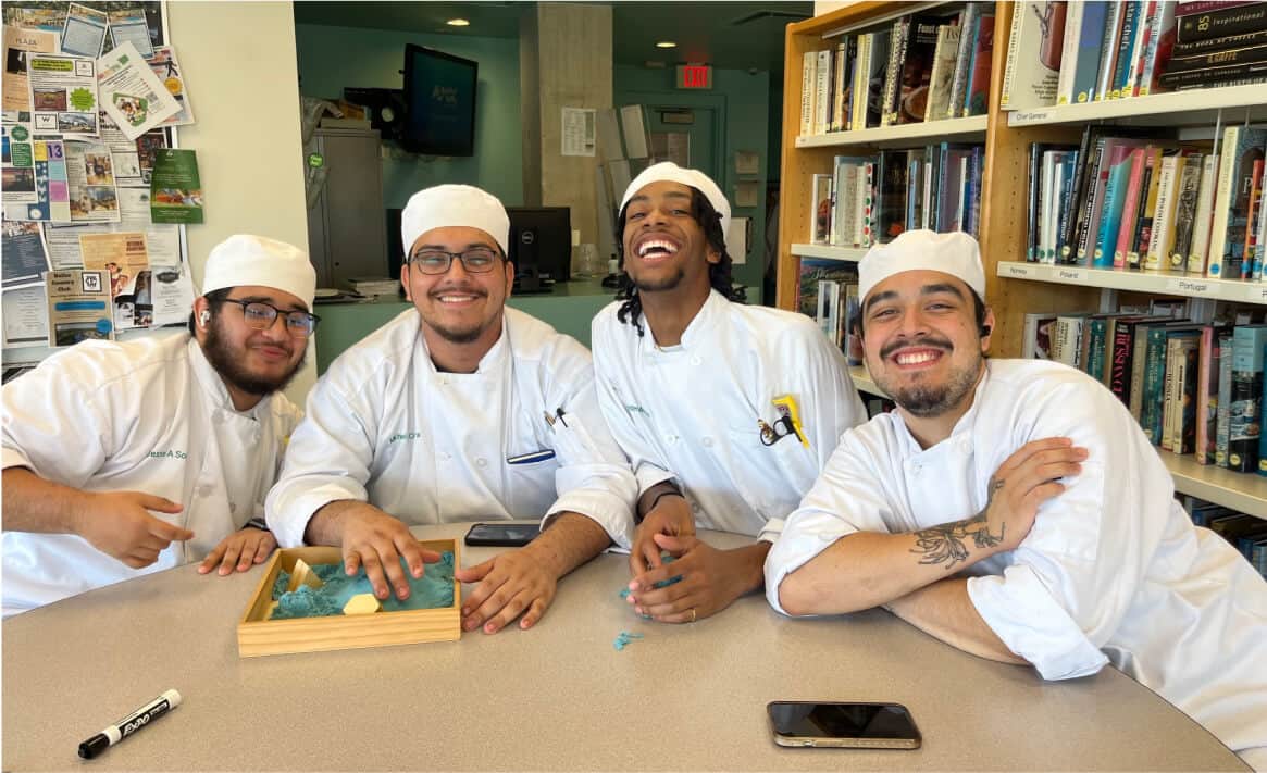 Four people dressed in white chef uniforms and hats are seated around a table in a room with bookshelves, embodying the vibrant brand of student life. They are smiling and engaged with a board game, while a smartphone and dry erase marker add to their dynamic interaction.