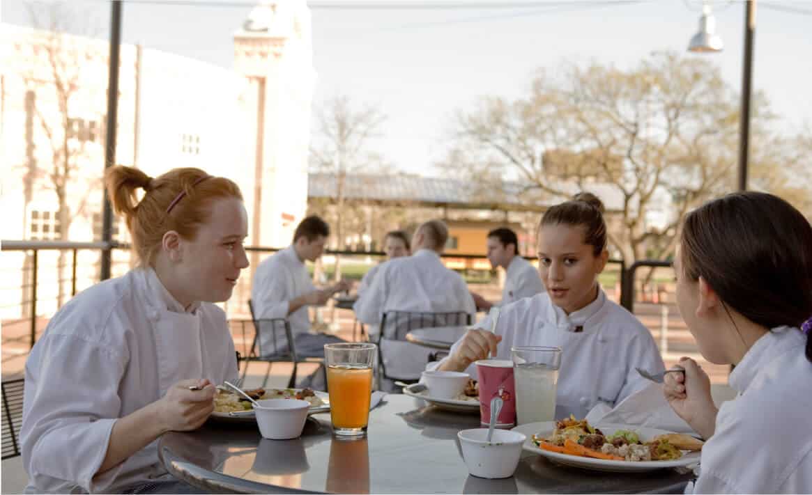 Three uniformed individuals, presumably chefs or culinary students, are seated at an outdoor table, enjoying a meal. The table is filled with plates of food and drinks. In the background, other similarly dressed people can be seen eating and engaging in conversation, embodying the essence of student life.